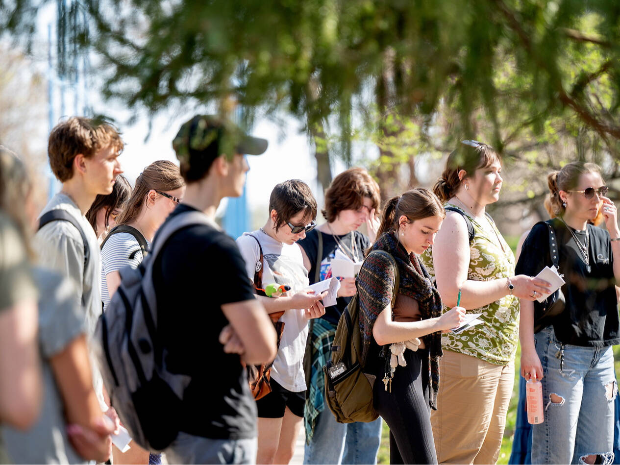 A group of young k-12 students stand outdoors under trees, some holding papers and writing, while others look around or use their phones.